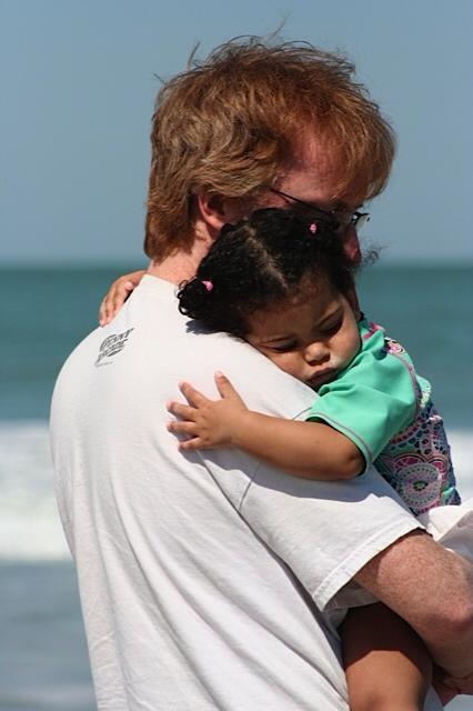 A toddler-age Rebecca snuggled up asleep against the shoulder of the author, who holds her in his arms as he gazes out to sea.