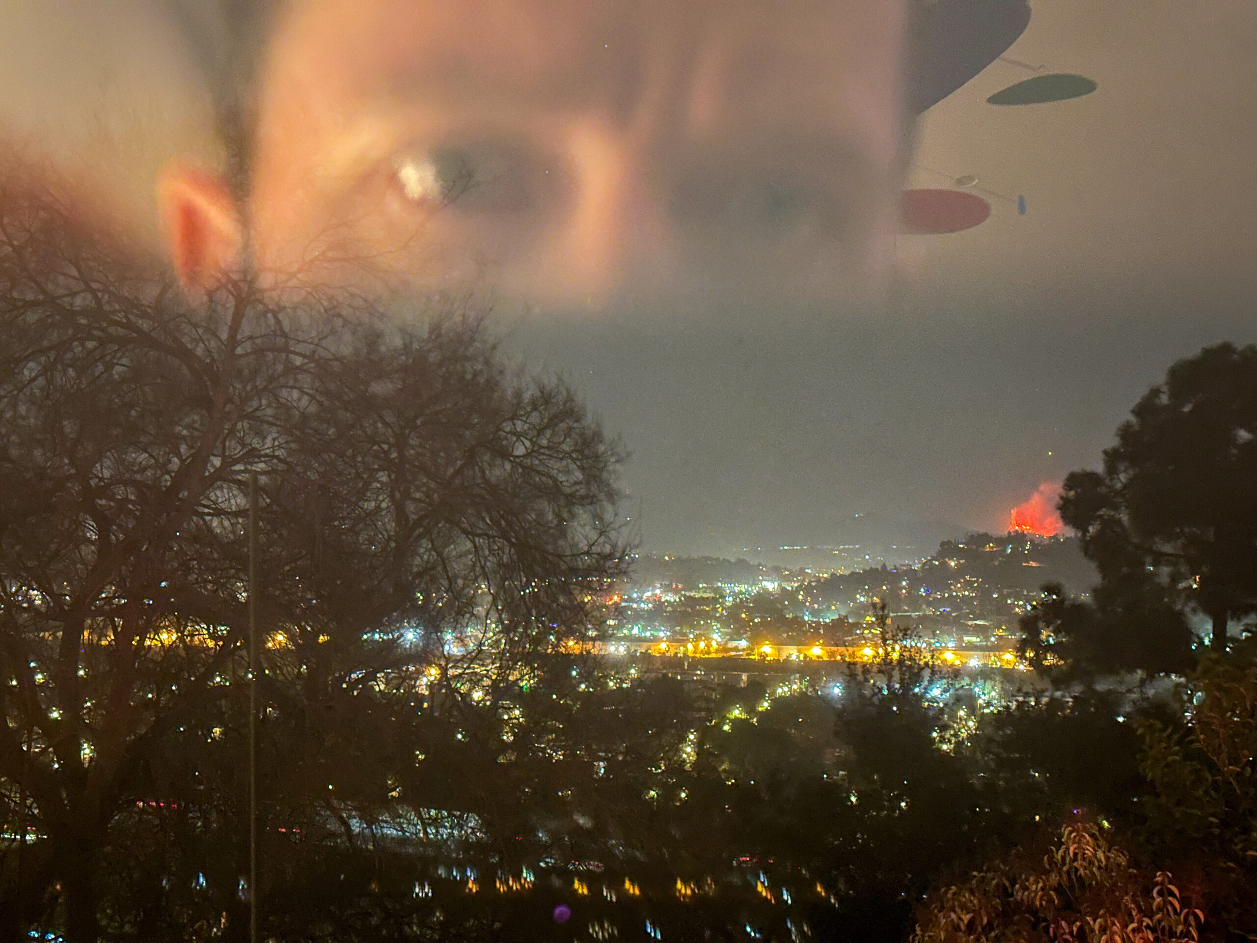 Nighttime view over Altadena with city lights below and a wildfire burning bright orange on a distant hillside; the faint reflection of a person’s face appears in the window glass in the foreground, watching as smoke and fire glow approach the neighborhood.