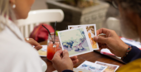 Two people sitting outdoors looking through old Polaroid photographs together, with one person holding up several photos to view.
